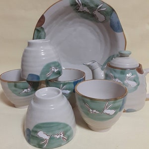 Op de afbeelding: Een witte keramische theeset met een groen en bruin ontwerp met konijnen. De set bevat een theepot, vier theekopjes en een serveerschaal.