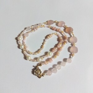 May include: A delicate necklace featuring a mix of pink gemstones, pearls, and small beads. The necklace has a gold-colored clasp and is arranged on a white surface. The gemstones vary in shape and size, creating a unique and elegant design.