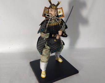 May include: A Japanese samurai doll wearing a black and gold armor with a sword. The doll is standing on a black base.