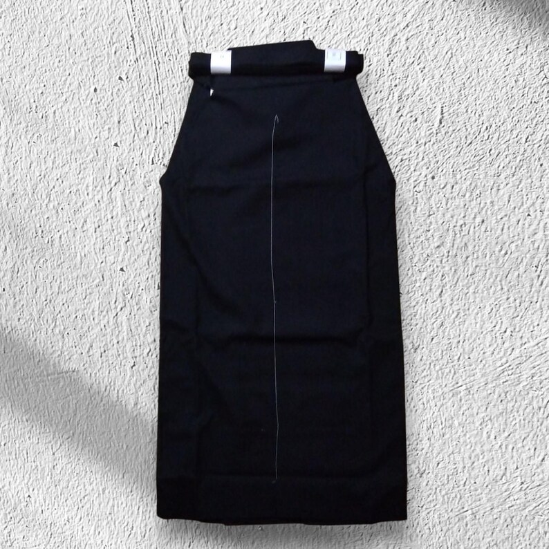 May include: A black hakama, a traditional Japanese garment, is displayed against a textured white background. The hakama features a wide, pleated design with a white seam running down the center and white accents at the top.