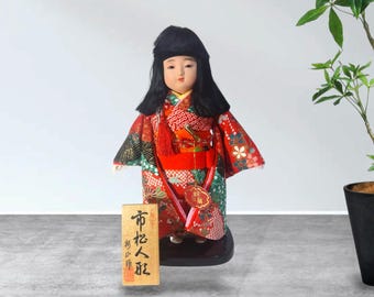 May include: A Japanese doll with black hair wearing a red kimono featuring intricate floral patterns in green, white, and gold. The doll stands on a black base next to a rectangular wooden plaque with black Japanese calligraphy and a red seal.