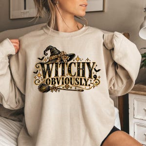 May include: A beige crewneck sweatshirt featuring a graphic print with the text "WITCHY OBVIOUSLY." The design includes a black witch hat with a leopard print bow, a broomstick, small bats, and gold star accents in a decorative layout.
