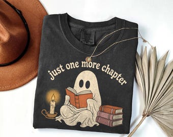 May include: A charcoal gray t-shirt featuring a graphic of a white ghost reading an orange book. The text "just one more chapter" is printed above the ghost. A lit candle in a holder is on the left, and a stack of three books is on the right.