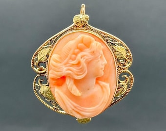 May include: Oval cameo pendant featuring a profile of a woman with flowing hair, carved in peach-colored material. The cameo is set in a gold filigree frame with gold leaf accents and a bail for hanging.