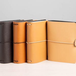 Op de afbeelding: Vier leren tijdschriften in bruin- en beigetinten. De tijdschriften zijn gesloten met een elastisch koord. Het eerste tijdschrift is donkerbruin met een textuur, gevolgd door een medium bruin en twee beige tijdschriften. De tijdschriften staan rechtop.