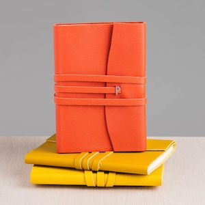 Puede incluir: Dos diarios encuadernados en cuero apilados uno encima del otro. El diario superior es naranja y tiene una correa de cuero con una letra 'P' plateada. El diario inferior es amarillo.