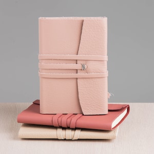 Può includere: Tre diari rilegati in pelle impilati uno sopra l'altro. Il diario superiore è di colore rosa chiaro con una chiusura argentata e due cinturini in pelle. Il diario intermedio è di colore rosa scuro e il diario inferiore è di colore beige chiaro.