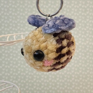 May include: A handmade, plush keychain featuring a round, fuzzy creature with a light yellow and brown body, black eyes, and a pink stitched mouth. It has blue, fluffy ears and a silver keyring.