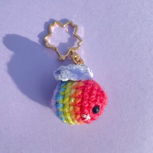May include: A handmade crochet keychain featuring a colorful, round creature with a gold-colored flower-shaped clasp. The creature is primarily red, with bands of yellow, green, and blue. It has small black eyes and a white stitched mouth.