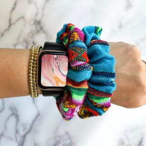 May include: A colorful woven fabric scrunchie in shades of blue, green, red, yellow, and pink. The scrunchie is worn on a wrist with a gold beaded bracelet and a black Apple Watch with a white and pink abstract background on the screen. The time on the watch is 10:09.
