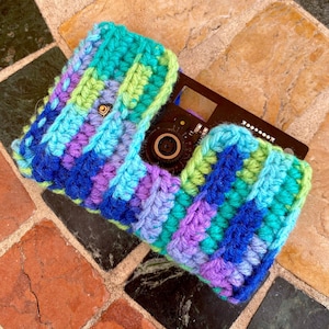 May include: A colorful, crocheted camera case in shades of blue, green, and purple. The case is rectangular with a flap closure and is designed to fit a small camera. The case is handmade and has a textured appearance.