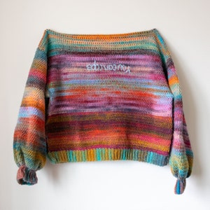 May include: A multi-coloured crochet sweater with a wide neckline and long sleeves. The sweater has a striped pattern in shades of orange, red, purple, blue, and green. The word "handmade" is embroidered on the front of the sweater.
