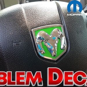 May include: A close-up of a black steering wheel with a silver and green Ram emblem. The emblem has the words "Mopar" and "Official Licensed Product" on it. The steering wheel also has a horn button and a control panel with buttons labelled "On/Off", "Cancel", "Set", and "Res".