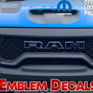 May include: A blue Ram pickup truck with a black grille and a chrome RAM logo. The truck has a black hood scoop and a Mopar logo on the front fender. The text "OFFICIAL LICENSED PRODUCT" is visible on the right side of the image. The text "EMBLEM DECALS" is visible at the bottom of the image.