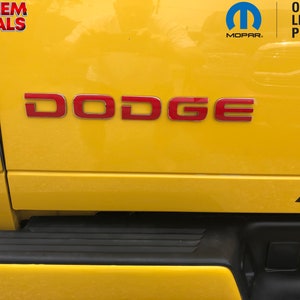 Peut inclure: Un pick-up Dodge Ram jaune avec un emblème Dodge rouge sur le hayon. Le camion a un pare-chocs noir et une plaque d'immatriculation noire. Le camion est garé sur une surface pavée.
