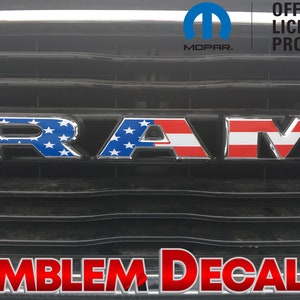 May include: A silver "RAM" emblem with a red, white, and blue American flag design. The emblem is on a black background with the text "OFFICIAL LICENSED PRODUCT" and "MOPAR" in white. The text "EMBLEM DECALS" is in red at the bottom of the image.