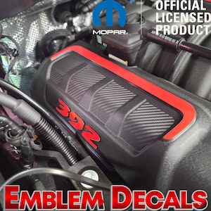 May include: Black engine cover with red trim and the number "392" in red. The cover has a carbon fiber pattern and the Mopar logo. The text "OFFICIAL LICENSED PRODUCT" is visible. The words "EMBLEM DECALS" are also visible.
