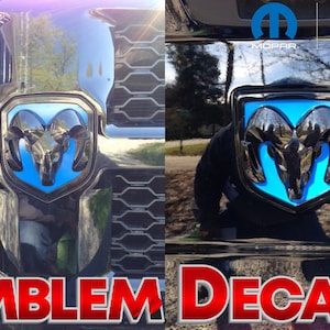Peut inclure: Deux autocollants d'emblème Ram bleus et noirs. Les autocollants sont des produits Mopar sous licence officielle. Le texte "EMBLEM DECALS" est affiché en rouge en bas de l'image.