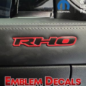 May include: Close-up of a black surface with a red and black "RHO" emblem. The emblem is outlined in red and has a black interior. The text "EMBLEM DECALS" is displayed in red at the bottom. The Mopar logo is visible.