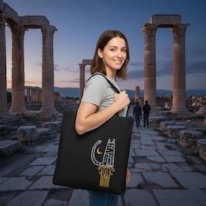 May include: Black tote bag with a white and gold cat design, including a crescent moon, perched on a decorative column. The bag has black straps. The design is a stylized illustration.