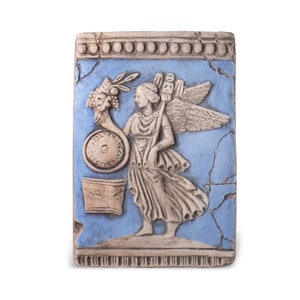 Puede incluir: Un azulejo de cerámica con fondo azul y una escultura en relieve blanca de una figura alada que sostiene un escudo y una rama de fruta. El azulejo tiene un borde decorativo de puntos blancos.