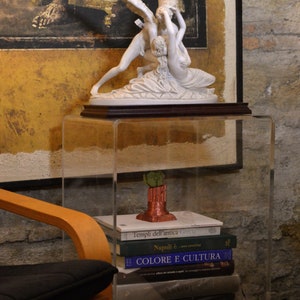 May include: A white porcelain sculpture of two angels on a wooden base, displayed on a clear acrylic shelf with a stack of books underneath. The books have Italian titles, including "Templi dell'antica Grecia" and "Colore e Cultura".