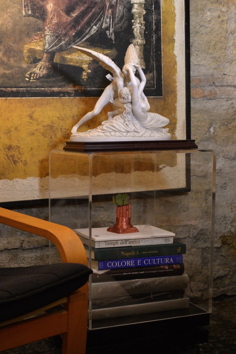May include: A white porcelain sculpture of two angels on a wooden base, displayed on a clear acrylic shelf with a stack of books underneath. The books have Italian titles, including "Templi dell'antica Grecia" and "Colore e Cultura".