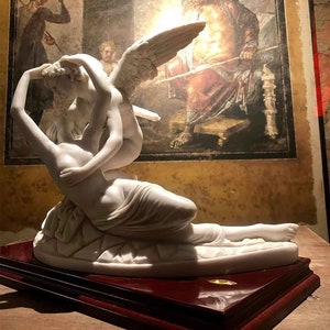 May include: A white marble sculpture of a winged figure embracing a reclining figure. The sculpture is on a dark wood base.