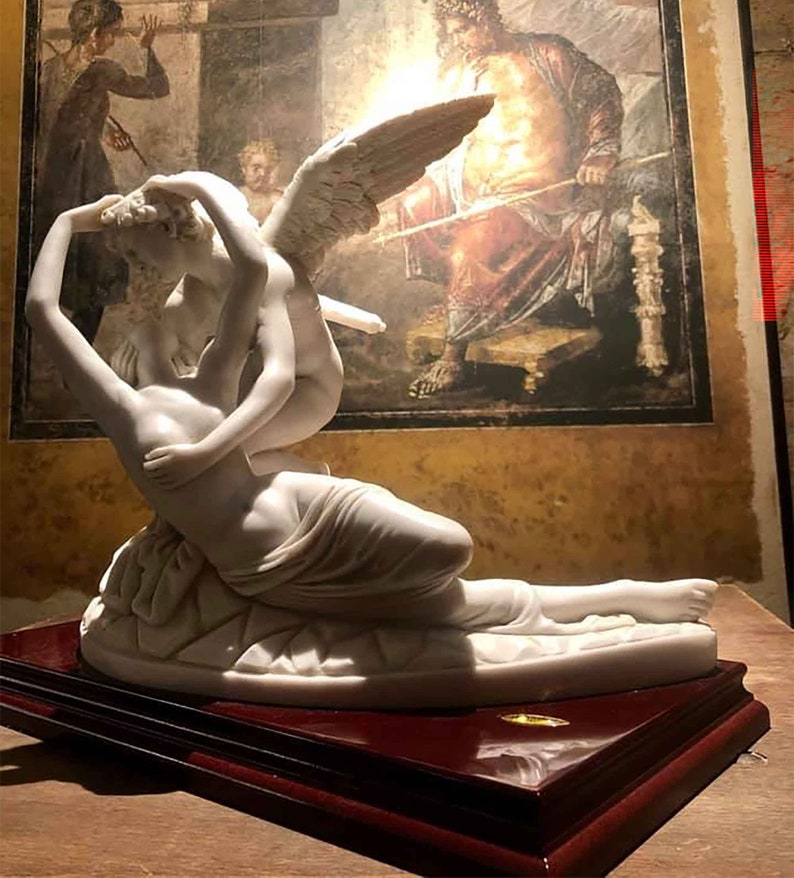 May include: A white marble sculpture of a winged figure embracing a reclining figure. The sculpture is on a dark wood base.