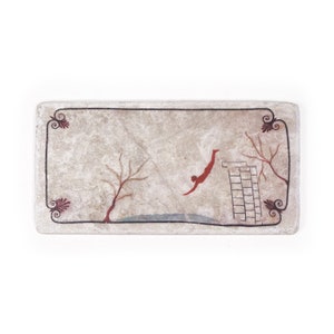 Op de afbeelding: Een rechthoekige tegel van steen met een geschilderd tafereel van een persoon die van een platform in het water duikt. De scène wordt omlijst door een decoratieve rand met gestileerde bloemmotieven in rood en zwart.