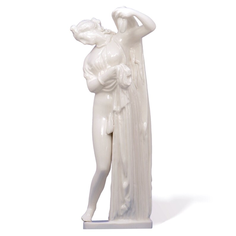 Puede incluir: Una escultura de porcelana blanca de una mujer de pie con los brazos en alto, sosteniendo una tela sobre su cabeza. La escultura es de estilo cl&aacute;sico.