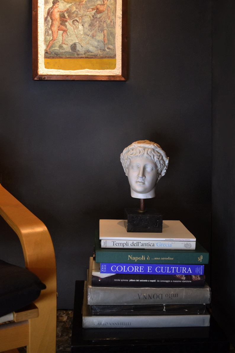 May include: A white plaster bust of a man with a laurel wreath on his head, sitting on a stack of books. The books have titles in Italian, including "Templi dell'antica Grecia" and "Napoli &egrave;... una cartolina".