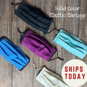 May include: Five face masks in different solid colors, including black, purple, blue, light blue, and cream. The masks are pleated and have elastic ear loops. The text "Solid Color Elastic Earloop" is written in white on a brown background. The text "SHIPS TODAY" is written in red on a white circle.