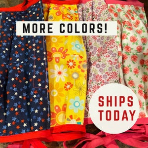 May include: Four fabric face masks with different patterns: blue with white stars, yellow with colorful flowers, pink with white flowers, and white with pink roses. The text "MORE COLORS!" is written in white on a black background. The text "SHIPS TODAY" is written in red on a white circle.