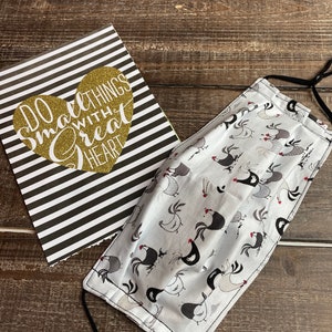 May include: A white face mask with a black and white rooster pattern. The mask has black elastic ear loops. The mask is lying on a wooden surface next to a card with the text "Do small things with a great heart".