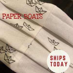May include: White fabric with a black and white pattern of paper boats. The fabric is folded in thirds. The text "PAPER BOATS" is printed in red at the top of the fabric. The text "SHIPS TODAY" is printed in red on a white circle at the bottom of the fabric.