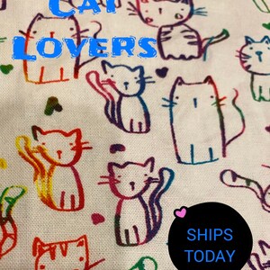 May include: White fabric with a colorful, hand-drawn pattern of cats. The text "CAT LOVERS" is written in blue letters across the top of the fabric. A black circle with the text "SHIPS TODAY" is in the lower right corner.