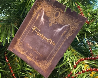 May include: A brown decorative ornament shaped like a book, with the title "Frankenstein" and author "Mary Shelley" in gold lettering. The ornament has a gold decorative design with lightning bolts and wings. The ornament is hanging on a Christmas tree.