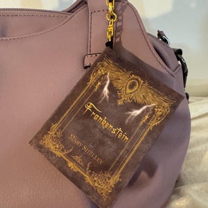 May include: A mauve handbag with a gold-coloured clasp and a decorative book charm. The book charm is a miniature version of "Frankenstein" by Mary Shelley, with gold lettering and lightning bolt accents.