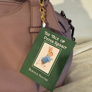 May include: A mauve handbag with a gold chain and a charm featuring a miniature book. The book cover is green with the title "The Tale of Peter Rabbit" and an illustration of a rabbit wearing a blue jacket.