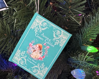 May include: A teal-colored ornament shaped like a miniature book, titled "Anne of Green Gables" by Lucy Maud Montgomery. The cover features a portrait of a young girl surrounded by flowers. The ornament hangs on a Christmas tree with colorful lights.
