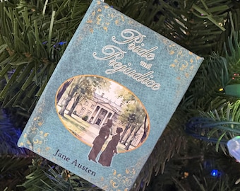 May include: A teal-colored ornament shaped like a book, featuring the title "Pride and Prejudice" and the author's name, Jane Austen. The cover illustration depicts two figures in front of a stately home. The ornament hangs from a white ribbon, nestled among Christmas tree branches.