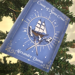 May include: A blue book cover with the title "The Count of Monte Cristo" and author "Alexandre Dumas". The cover features a ship sailing on the sea, crossed swords, and decorative swirls. The book is displayed on a Christmas tree.