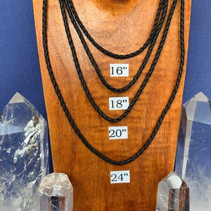 May include: Four black braided leather cords of varying lengths, 16 inches, 18 inches, 20 inches, and 24 inches, displayed on a wooden mannequin. The cords are shown against a blue background with two clear quartz crystals on either side of the mannequin.