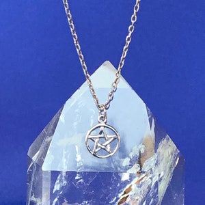 May include: A silver chain necklace with a pentagram pendant. The pendant is a five-pointed star with a circle around it. The necklace is displayed on a clear crystal.