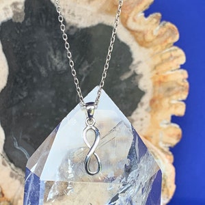 May include: A silver infinity symbol pendant necklace. The pendant is a figure eight shape, hanging from a fine chain. The necklace is displayed on a clear crystal with a blue backdrop.