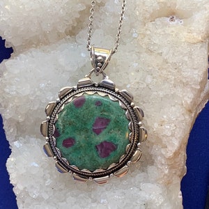 Può includere: Una collana con catena d'argento con un ciondolo rotondo che presenta una pietra preziosa verde e rosa incastonata in una cornice d'argento. Il ciondolo ha un bordo decorato a festoni.