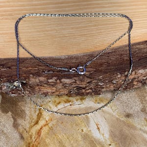May include: A silver-colored necklace with a delicate chain design. The necklace is displayed on a wooden surface, showcasing its intricate details and the clasp. The chain is made of small, rectangular links.