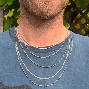 May include: A close-up of a man wearing a blue t-shirt with a silver chain necklace. The chain is layered in different lengths, creating a stylish and modern look.