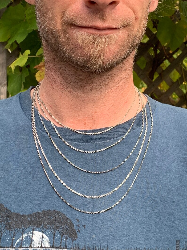 May include: A close-up of a man wearing a blue t-shirt with a silver chain necklace. The chain is layered in different lengths, creating a stylish and modern look.